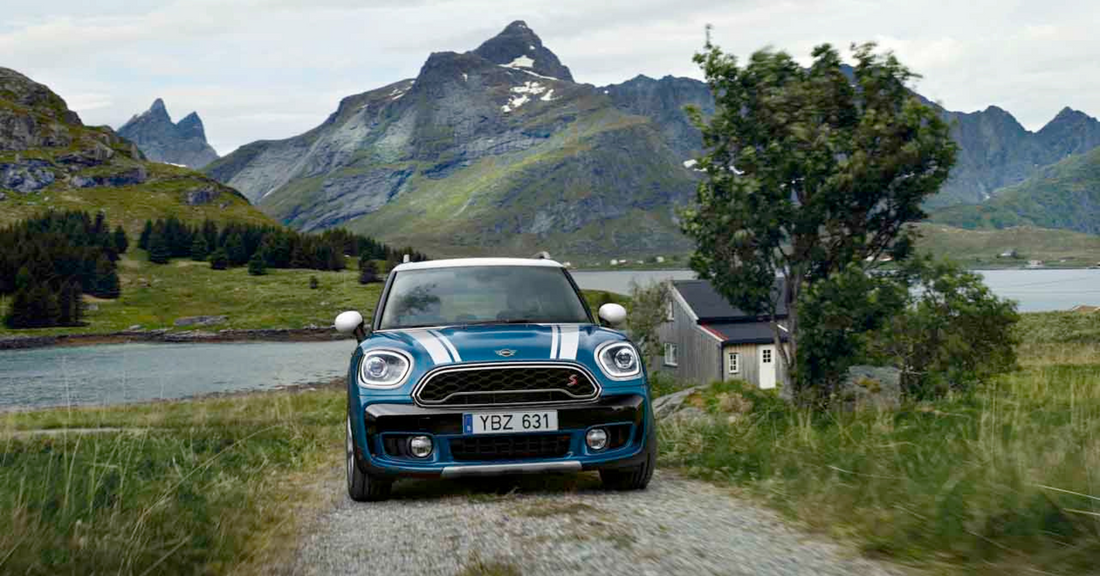 Blue MINI Cooper driving on a gravel road with scenic mountains and lake in the background